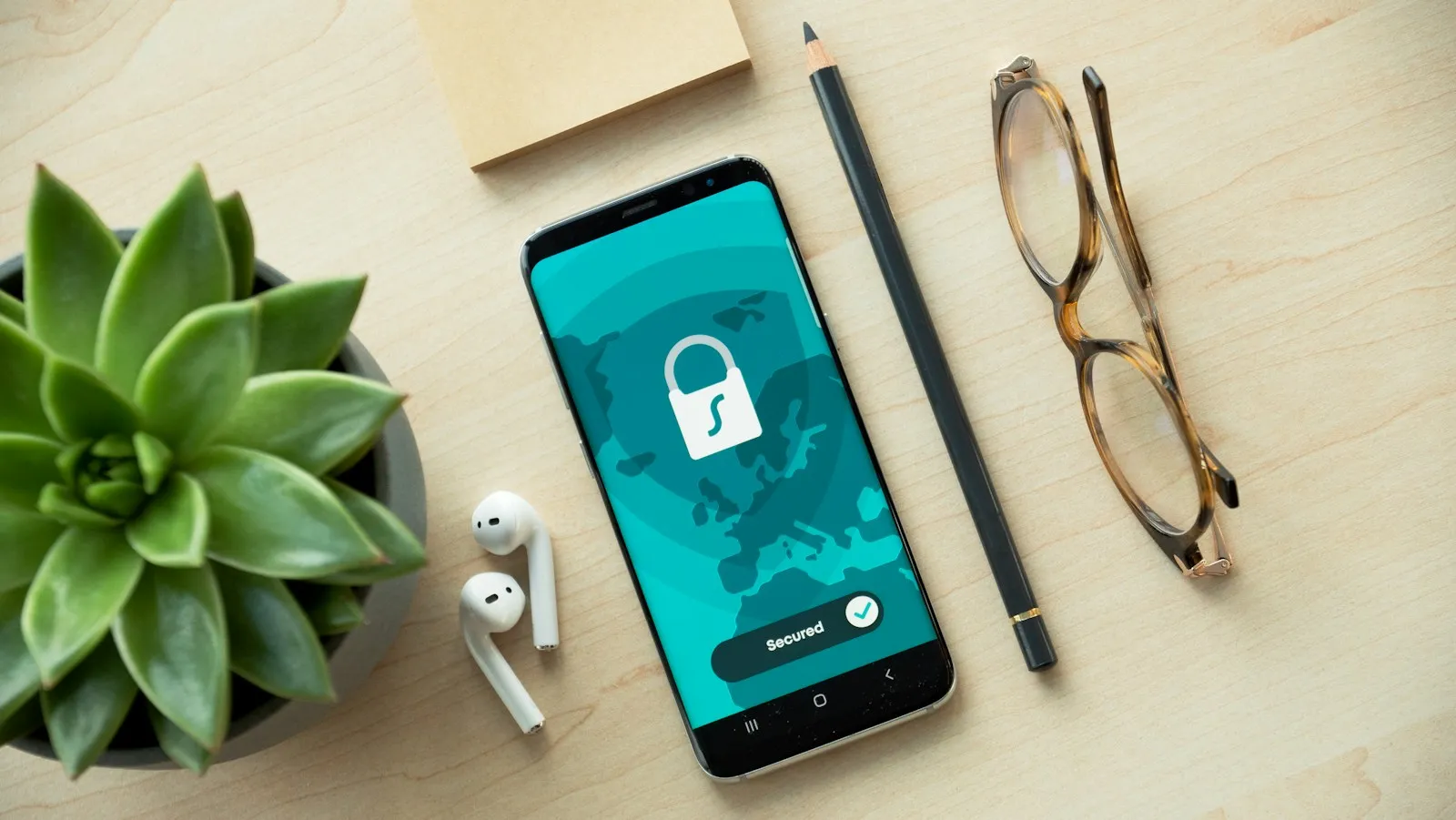 Celular com um cadeado na tela sobre uma mesa, ao lado de óculos, fones de ouvido e uma planta